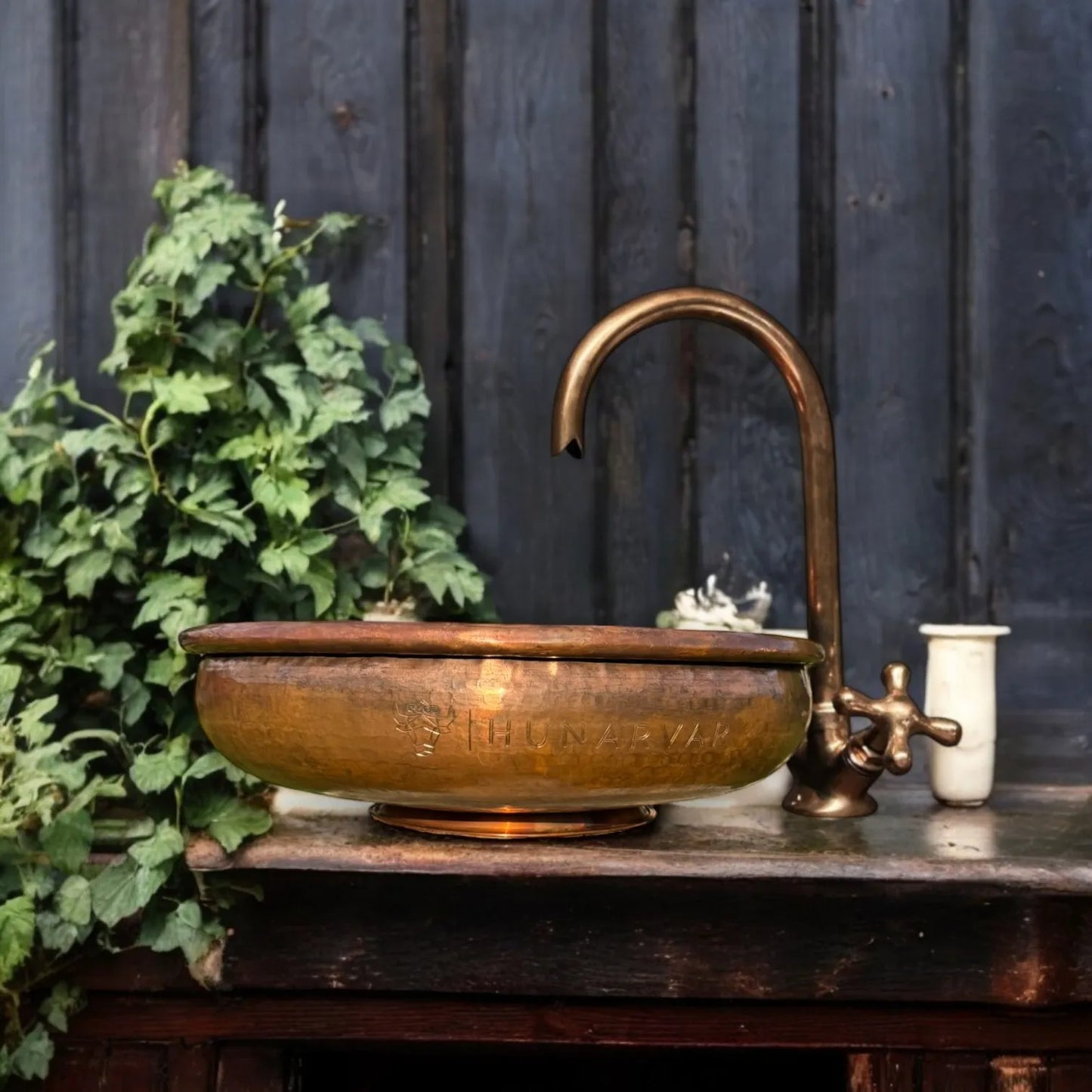 Gedengeltes Kupferwaschbecken mit passendem Wasserhahn auf einer rustikalen Holzplatte, im Vintage-Stil, vor einer dunklen Holzwand und mit grünen Pflanzen als Dekoration.
