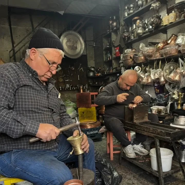 Zwei türkische Handwerker bearbeiten Kupfer in einer traditionellen Werkstatt, umgeben von Metallwaren und Werkzeugen.
