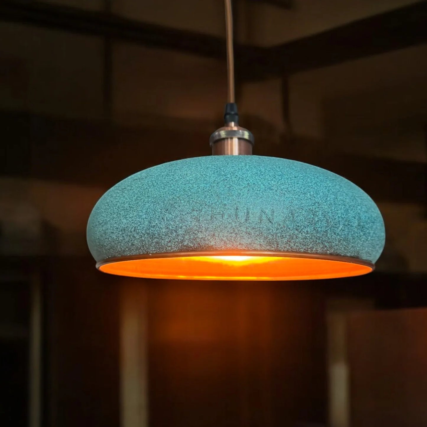 Türkis-blaue Kupfer-Pendelleuchte, von oben hängend, mit warmem Lichtschein vor unscharfem, braunem Hintergrund.
