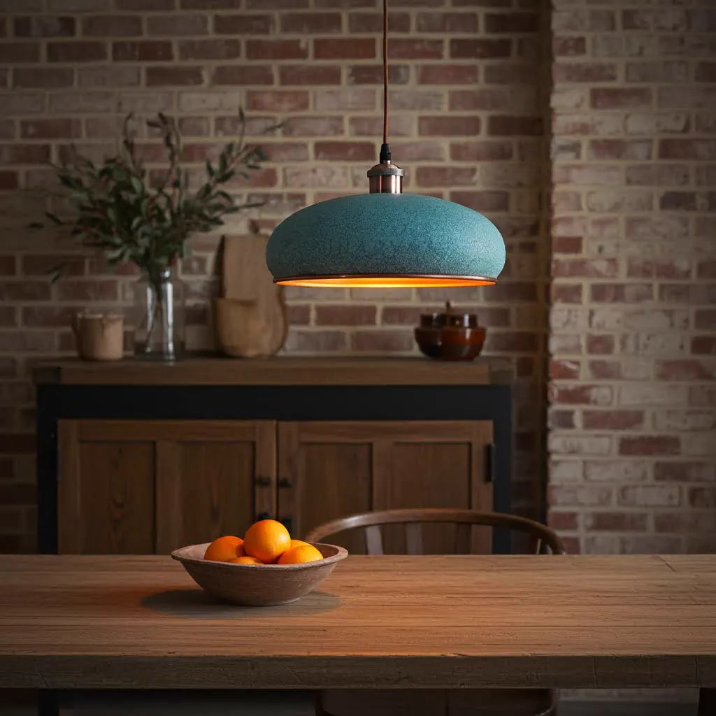 Essbereich mit Holztisch, Schale mit Orangen, türkisfarbener Kupfer Pendelleuchte und rustikaler Ziegelwand. Holzkommode mit Pflanze im Hintergrund.
