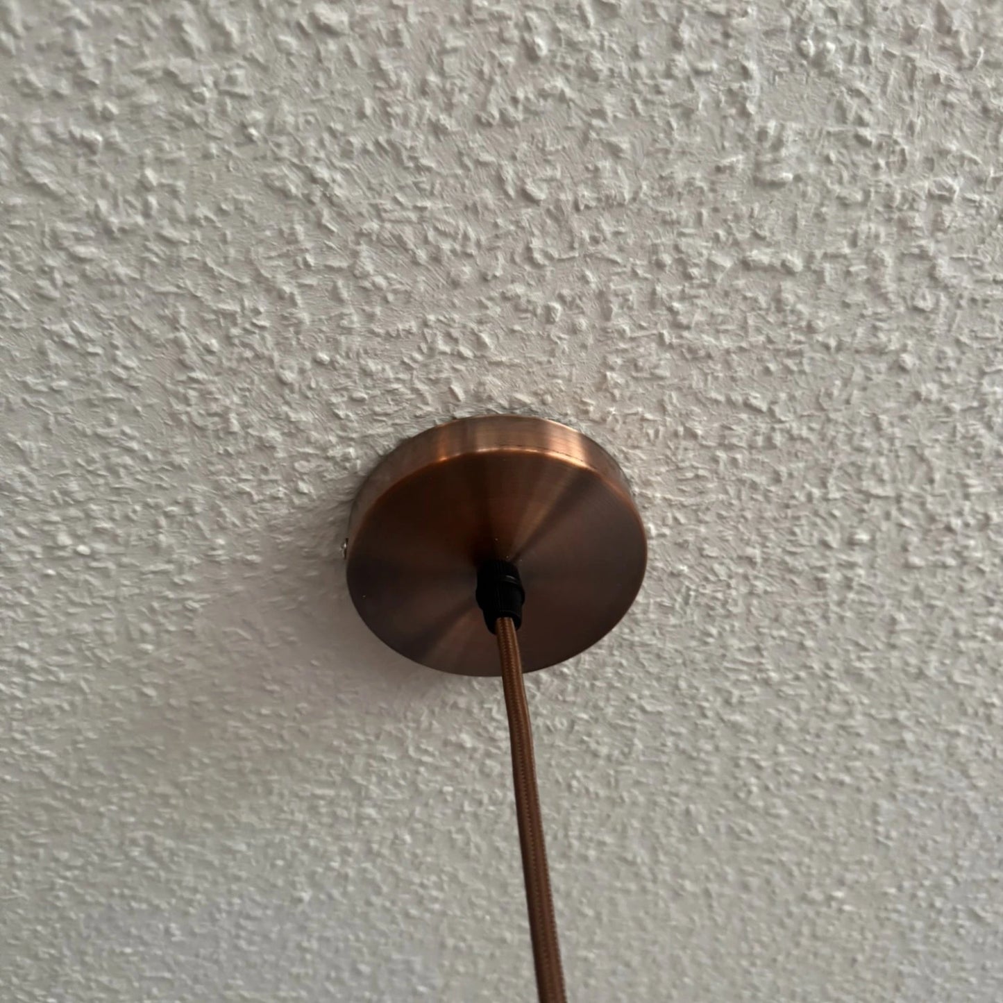 Kupferfarbene Deckenrosette einer Pendelleuchte, montiert an einer weißen Zimmerdecke, mit textilummanteltem Kabel.
