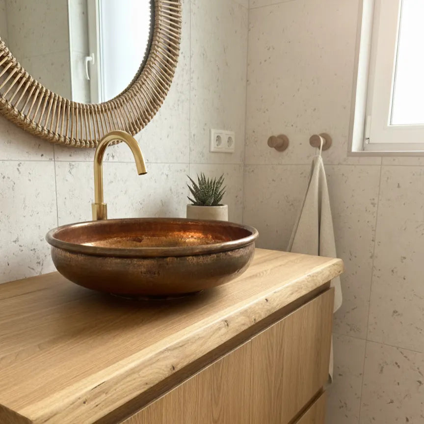 Handgefertigtes Waschbecken aus Kupfer auf einer massiven Holzplatte in einem modernen Badezimmer mit goldener Armatur und natürlicher Lichtstimmung
