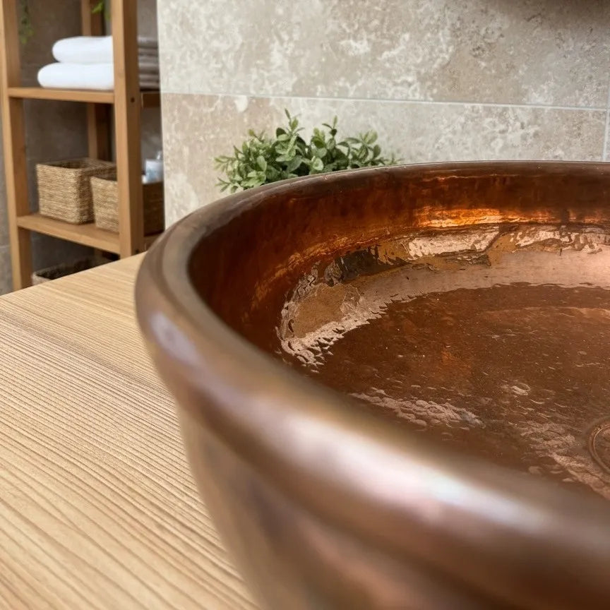 Detailaufnahme eines handgefertigten Kupferwaschbeckens auf einer Holzplatte in einem modernen Badezimmer mit Natursteinwand und dezenten Pflanzenakzenten