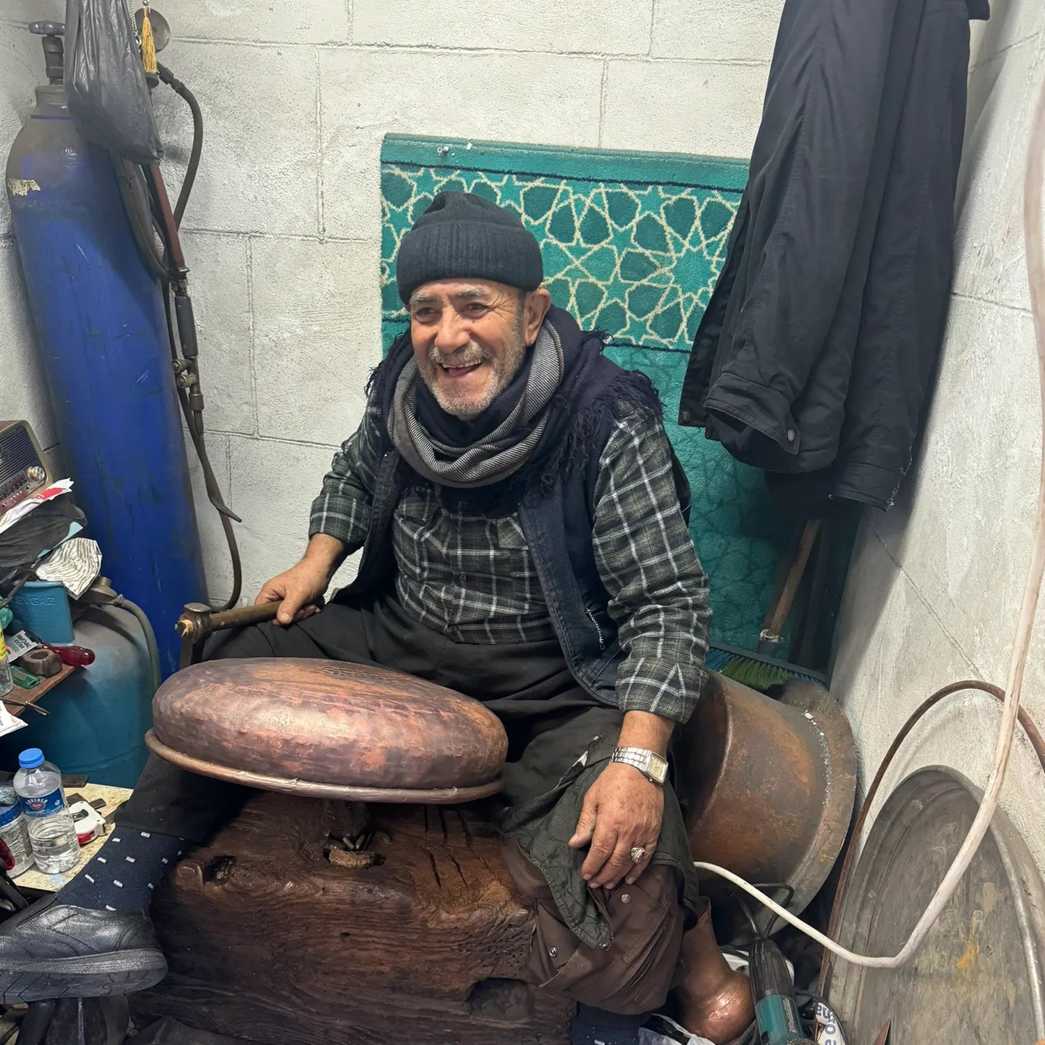 Älterer Mann sitzt lächelnd in einer Werkstatt, bearbeitet ein Kupferwaschbecken auf einem Holzblock, mit Werkzeugen im Hintergrund.